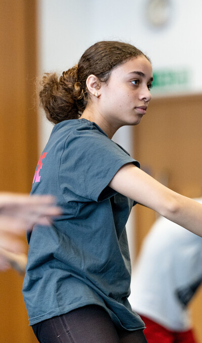 A dancer with brown hair, wearing a grey t-shirt has their arms in a curved position in front of their chest. They are looking forwards towards the mirrors in the studio