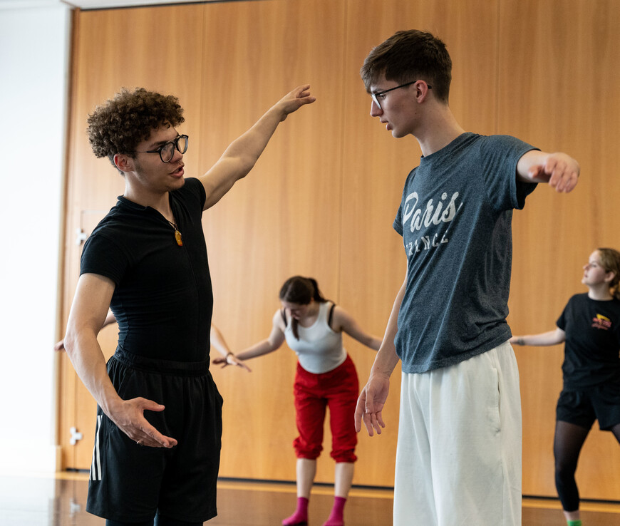 Two young students wearing glasses and dance clothing are working together. They have their arms outstretched to the side and are speaking to each other. In the background there are three other dancers