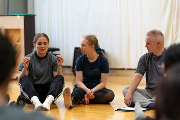 Three people are sitting on the floor in a discussion, one is the teacher Phil Hill. One student has their fingertips and thumbs touching and is mid talking, the other is looking towards them and appears to be listening.