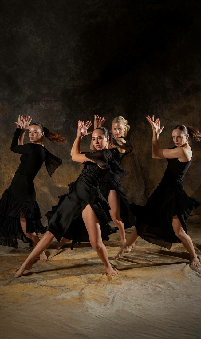 Four dancers in the same dynamic twisted position, hands in an expressive position near their faces. They are against a mottled fabric background.