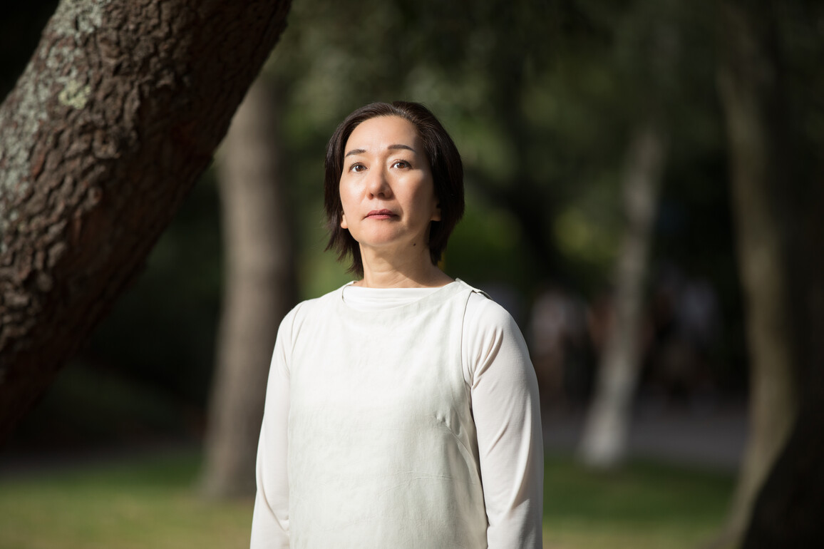 a lady is standing in a garden wearing a white top, looking just above at the camera. She has short brown hair.