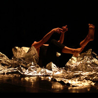 Image of pieces of fragments of mirrored card on the floor. Some pieces are more crumpled than others. A body is on their back with their legs up in the air. The background and floor are black.