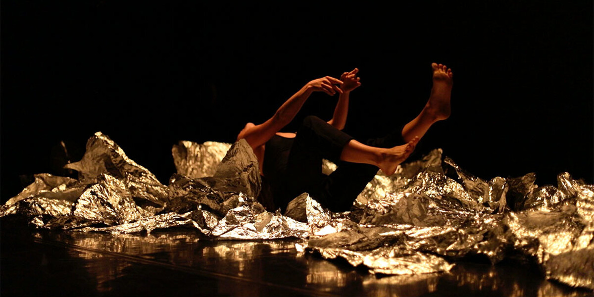 Image of pieces of fragments of mirrored card on the floor. Some pieces are more crumpled than others. A body is on their back with their legs up in the air. The background and floor are black.