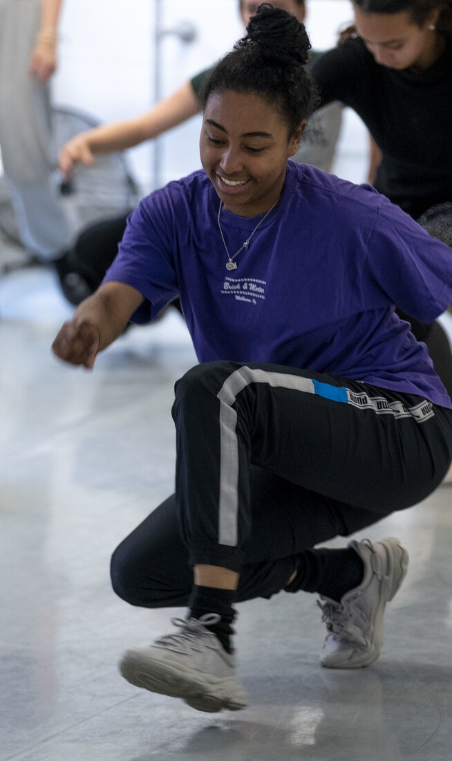 A teenager wearing a purple t-shirt and tracksuit bottoms in a dance studio crouching down and walking, there are several other teenagers in the studio behind them doing the same thing.