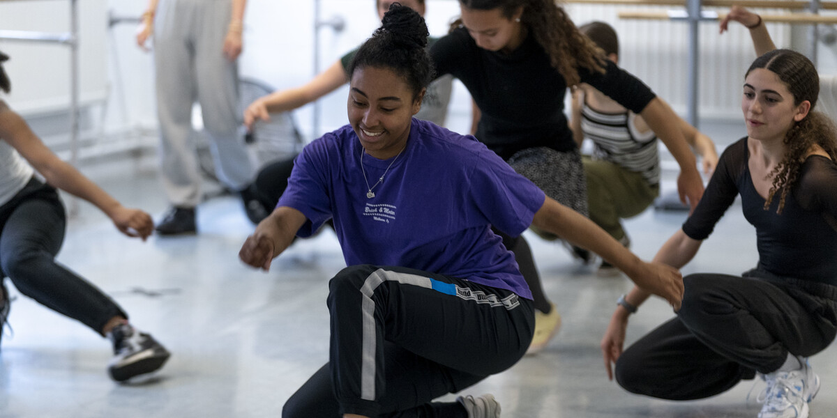 A teenager wearing a purple t-shirt and tracksuit bottoms in a dance studio crouching down and walking, there are several other teenagers in the studio behind them doing the same thing.