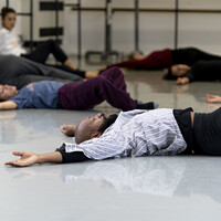 Contemporary teacher Gerrard Martin lies on the dance studio floor alongside some participants. Their arms are raised above their head and rest on the floor, and both knees are bent. They wear a variety of loose fitting athletic clothing.