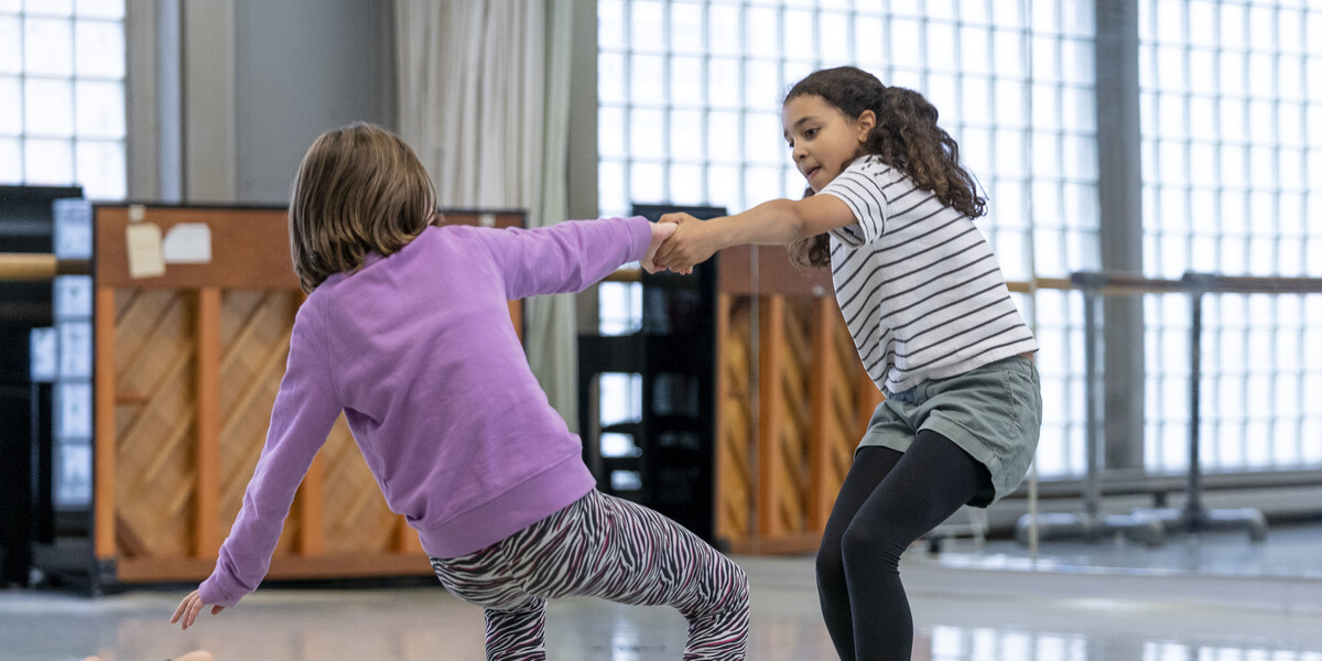 2 children in a dance studio wearing exercise clothes. They are holding hands and leading backwards.