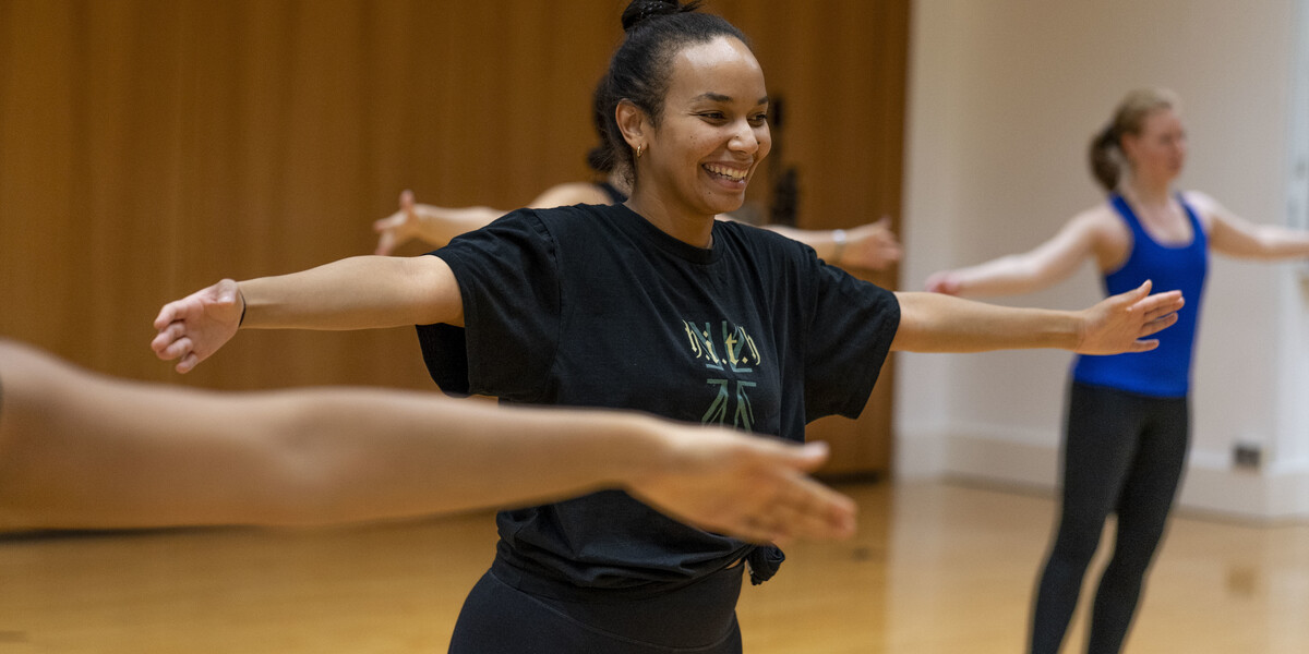 in a dance class a woman is standing with her arms extended by her side in a T shape, she is smiling and wearing a black t-shirt and black trousers. We see a few people behind her too.
