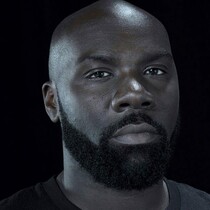 Portrait of Michael 'Mikey' J Asante, a black man with a beard. He is looking at the camera and is on a black background.