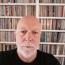 Portrait of Derek Hicks, a white man with a white beard and wearing a black t-shirt standing in front of a library of CDs