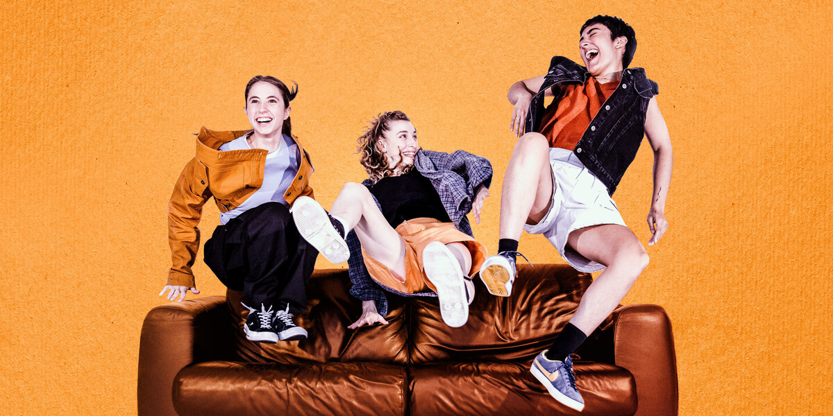 Three dancers leap up onto a brown leather sofa, smiling big warm smiles at each other, clearly they are close friends. Their outfits are all 90s inspired in shades of orange and red. The background of the image is an orange, textured like paper.