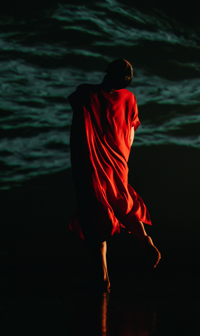 A figure draped in a red cloth, facing away from the camera towards the dark outline of a natural landscape in the background.