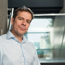 Eddie Nixon, Artistic Director of The Place is standing by a meshed metal wall in a staircase. He is a white man with dark hair, is wearing a blue shirt and is smiling.