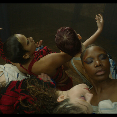 Image is of five dancers resting on each other with their arms raised. Their eyes are closed. They are wearing red and white costumes with colourful makeup and hair slicked back.