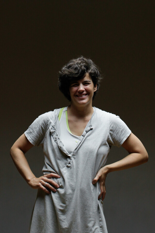 A woman wearing a baggy white t-shirt with short dark hair smiles at the camera with her hands on her hips. There is a dark background behind her