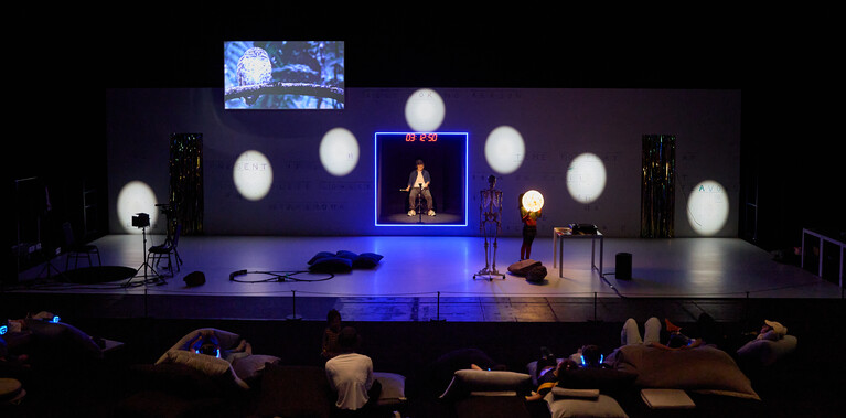 A photo of a stage with child performers across it, with a screen, props and light installations on stage with them