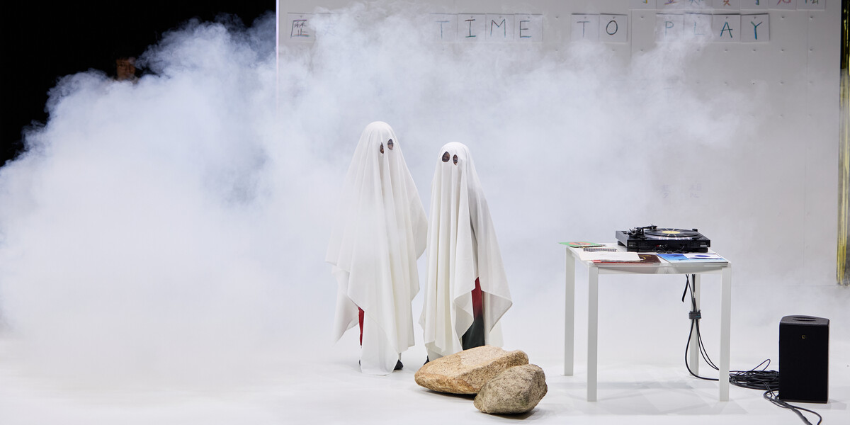 A photo of two child performers dressed as ghosts on a stage, wearing white sheets. A record player and stage smoke surrounds them.