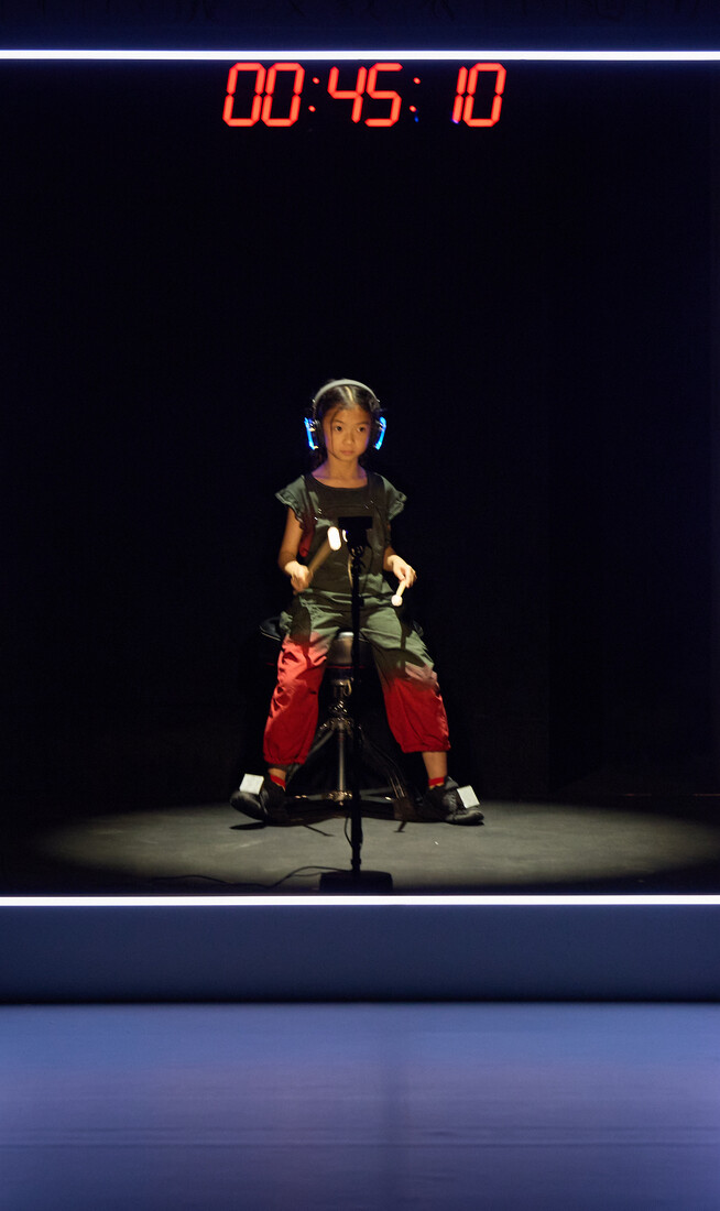 A photo of a child performer sat in a box with headphones and motion sensor drum kit. A digital clock with red lights is above them.