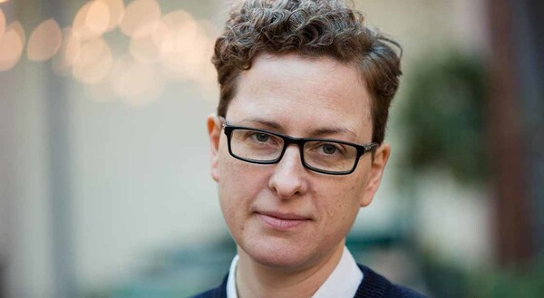 A woman with short hair, a soft smile and glasses looking directly into the camera, the background is blurry and her face is in focus.