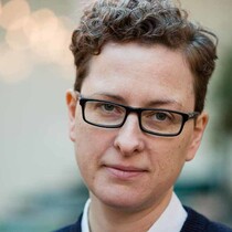 A woman with short hair, a soft smile and glasses looking directly into the camera, the background is blurry and her face is in focus.