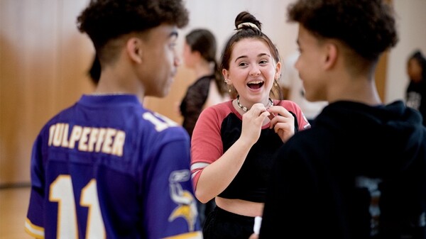 Two male students have their backs to the camera, looking at each other. One is wearing a purple sports t-shirt, the other a black hoodie. We see a young lady laughing between them. She is facing the camera and holding her necklace as she is looking at the young man on the left of the photo.