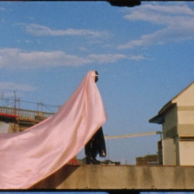 A dancer stands in the centre of the screen on a ledge. There is a bridge in the background. A long pink cloak fills the left side of the screen. The image has a retro vibe.