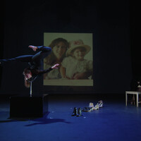 Image of a stage. A old photograph of a mother and daughter is suspended in the background. In the foreground on the right-hand side, Hannah Finn is on a black box, balancing in a position. Her body is contorted. On the left hand side are shoes and a small table.