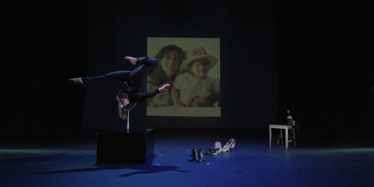 Image of a stage. A old photograph of a mother and daughter is suspended in the background. In the foreground on the right-hand side, Hannah Finn is on a black box, balancing in a position. Her body is contorted. On the left hand side are shoes and a small table.