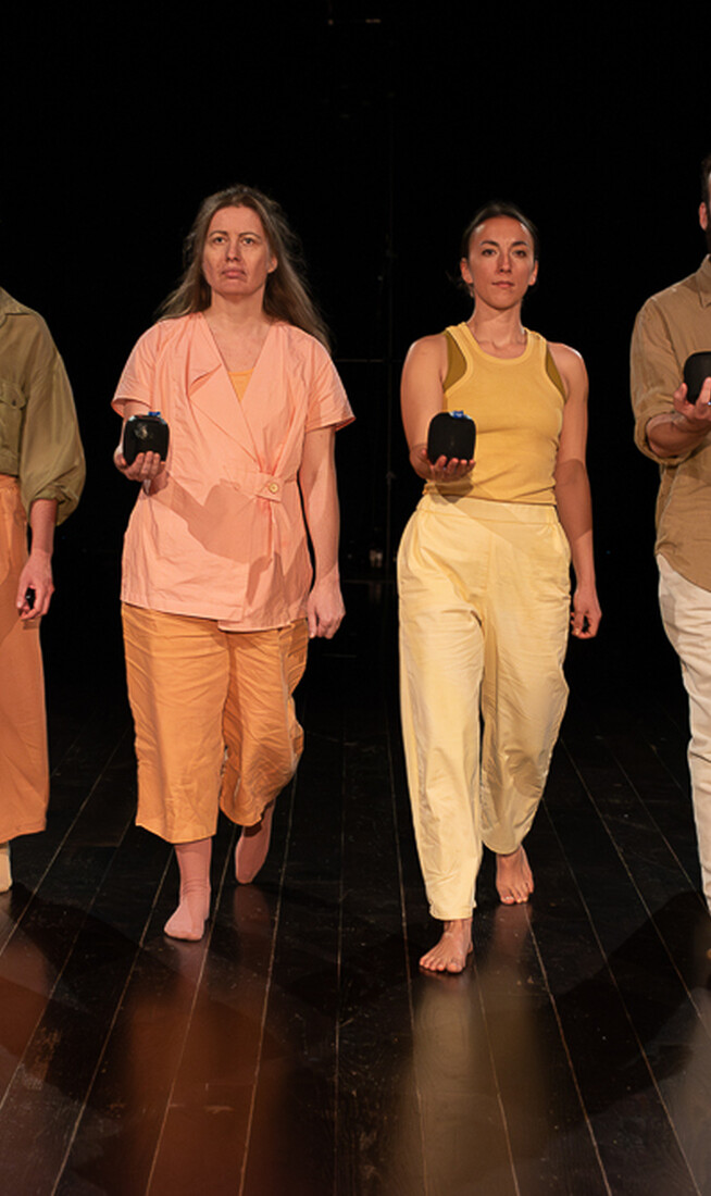 Four performers wearing soft pastel tones walk purposefully towards the camera in unison, each with one arm extended in front of them, portable bluetooth speakers in the palm of their hands.