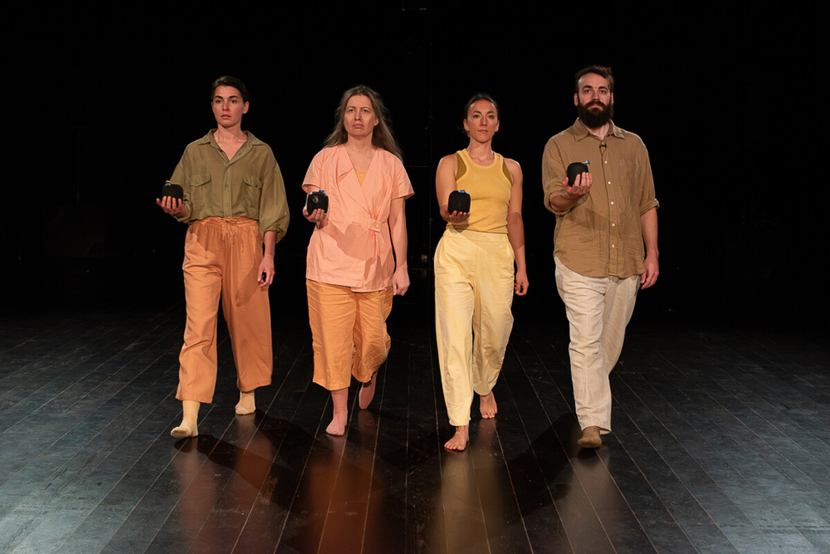 Four performers wearing soft pastel tones walk purposefully towards the camera in unison, each with one arm extended in front of them, portable bluetooth speakers in the palm of their hands.