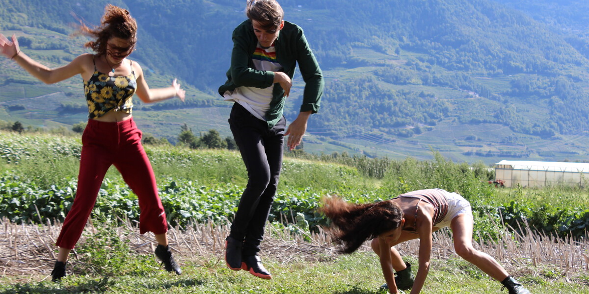 Image of three dancers dancing on green grass in the foreground. In the background are mountains
