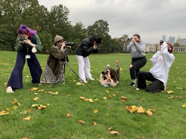 six students are standing or lying on grass in Greenwich Park, they have tripods and camera