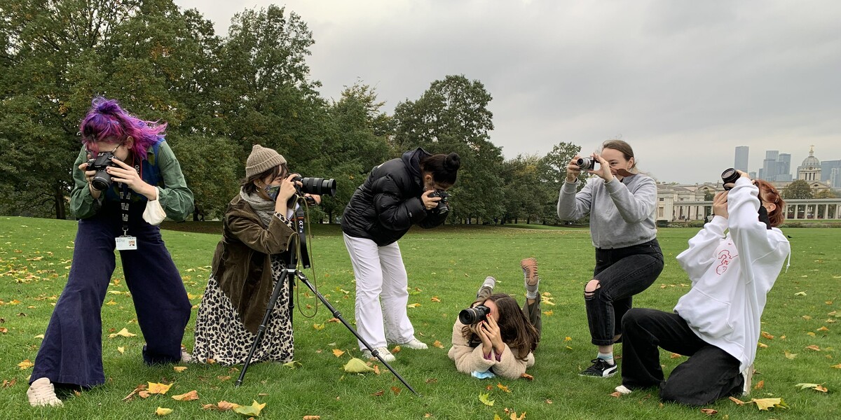 six students are standing or lying on grass in Greenwich Park, they have tripods and camera