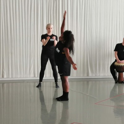 2 people in a dance studio, 1 is standing with the arms outstretched and the other appears to be directing them. There is a musician with a drum in the background.
