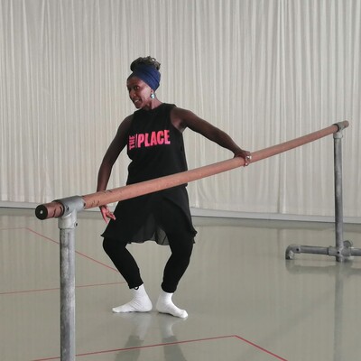 Ballet teacher in a The Place logo black t-shirt and leggings, they are bending at a ballet barre.