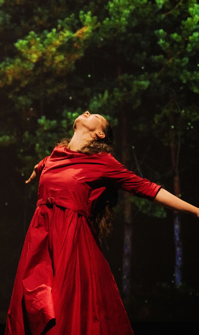 A woman wearing a red dress leaning backwards. Trees are on a screen in the background behind her.
