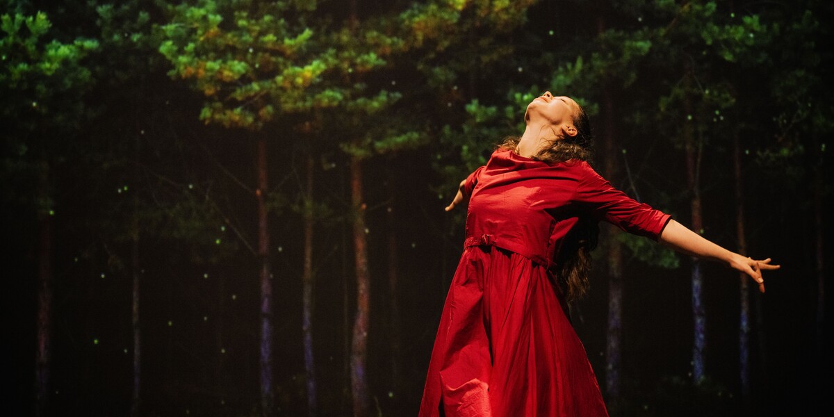 A woman wearing a red dress leaning backwards. Trees are on a screen in the background behind her.