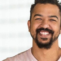 A headshot of a young man with a big smile and a goaty beard and short brown hair. He is wearing a pink t-shirt and is in front of a blurred window.