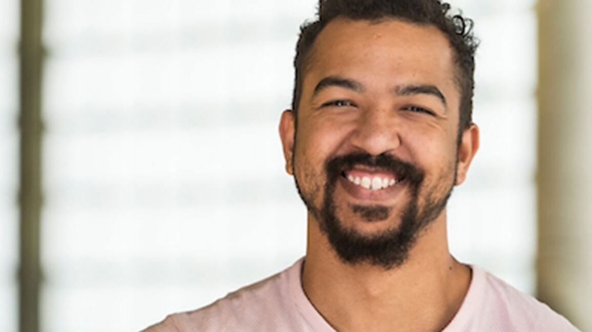 A headshot of a young man with a big smile and a goaty beard and short brown hair. He is wearing a pink t-shirt and is in front of a blurred window.