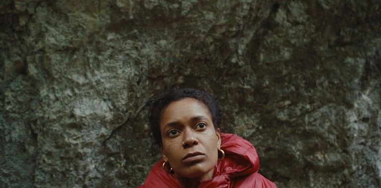 A performer stands in front of a large rock. You can only see their shoulders and head. They are wearing a red puffer jacket, gold hoop earrings and two nose rings. They are looking upwards and slightly concerned.
