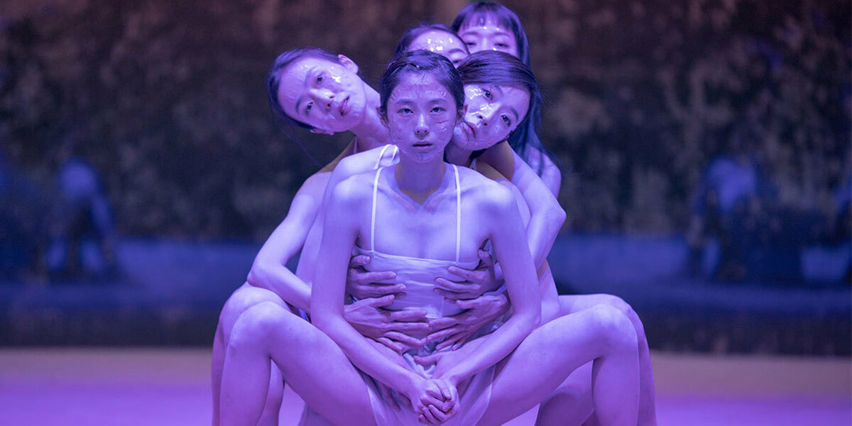 5 people sit on stage one behind the other. They are white vests and shorts. There is a purple hue on them.