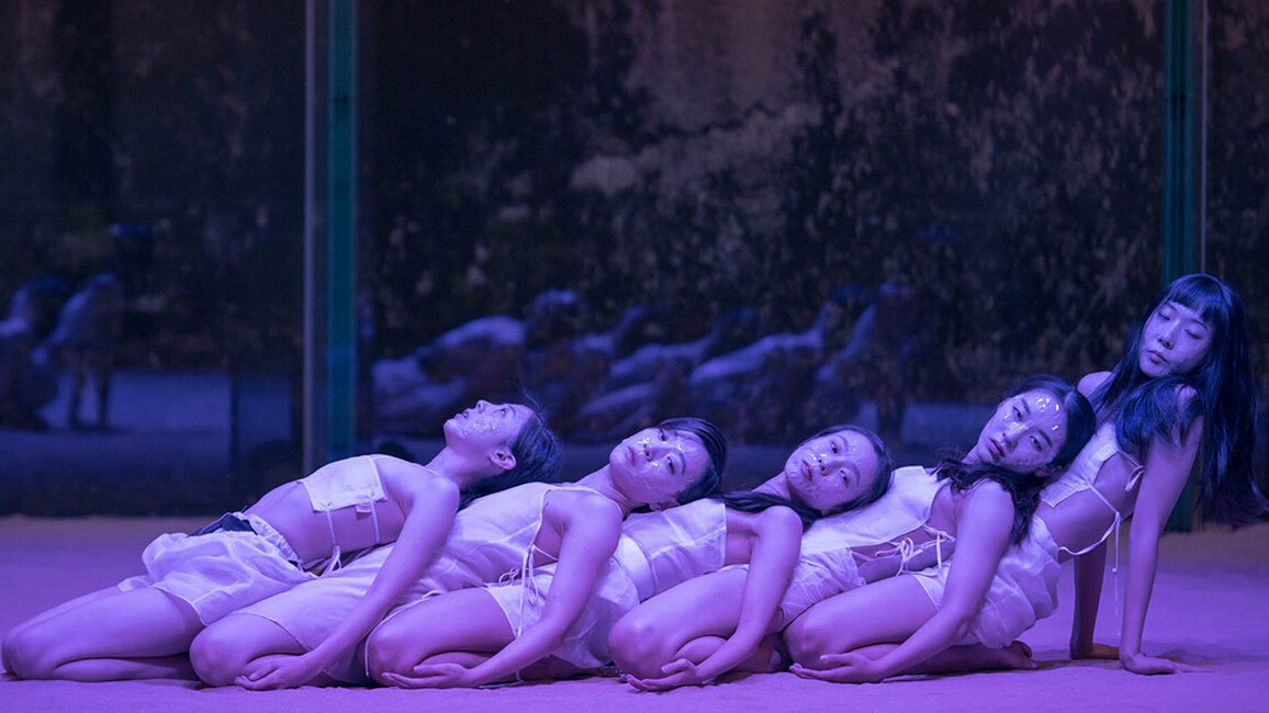 Five women basked in blue light and wearing white strappy costumes lie back on each other in a row.