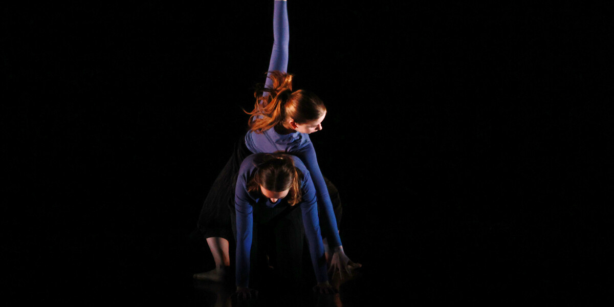An image of two dancers from Thrive Youth Dance Company creating a shape together, one of them in all fours position and another connected through their back and reaching up with their arm.