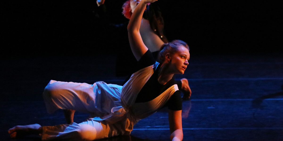 Image of Isis Clunie on a stage. Stage is darkly lit. Isis is wearing a blue shirt with white trousers. Isis lies on the floor with their left arm supporting them. Their right hand is raised up above them.