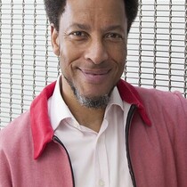 Image of Kenneth Olumuyiwa Tharp. Kenneth is a black man and wears a dark pink jacket with a lighter pink shirt underneath.