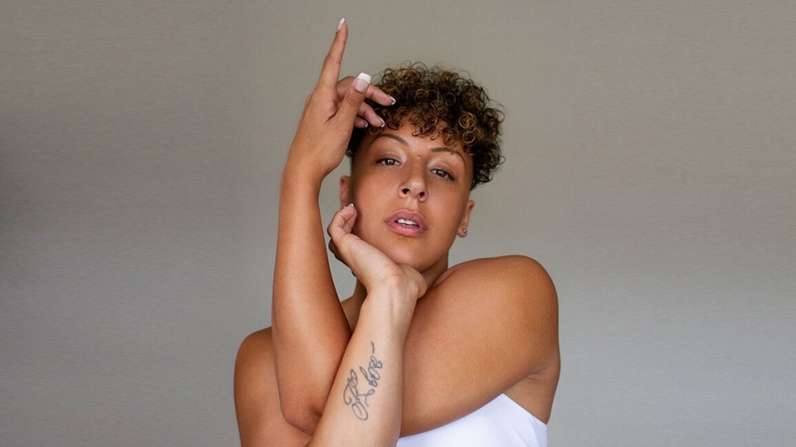 a performer is staring at the camera with their arms wrapped around each other in a voguing kind of pose. They are wearing a white sleeveless t-shirt