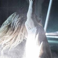 an abstract image of a performer with long blond hair dressed in a white dress looking away from the camera. There is a beam of light on her back and the background looks like a wearehouse.