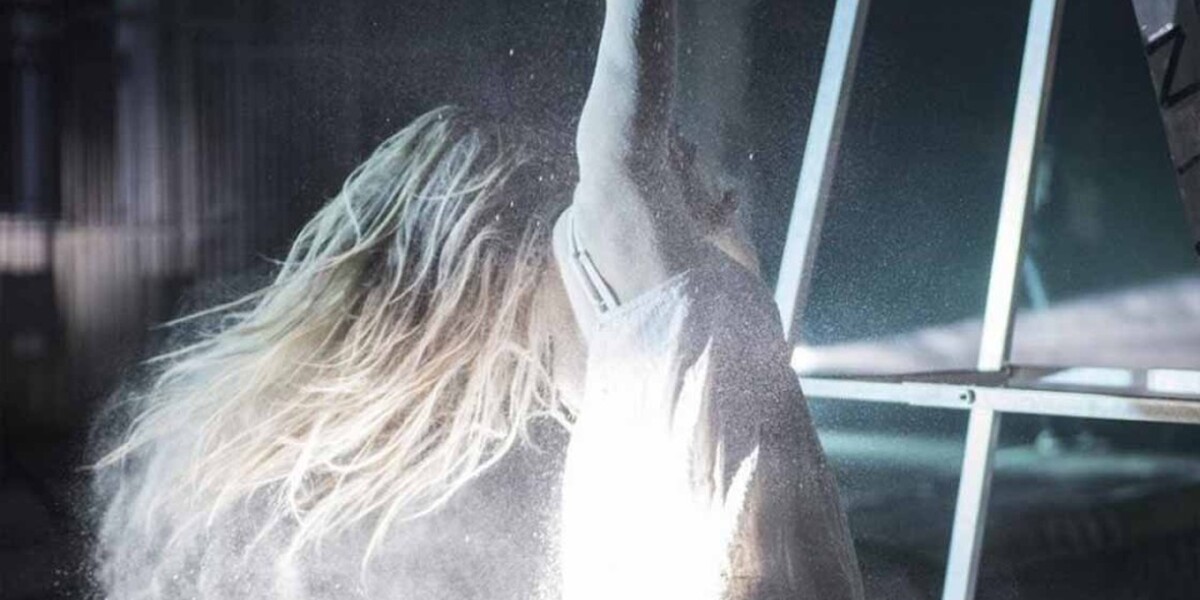 an abstract image of a performer with long blond hair dressed in a white dress looking away from the camera. There is a beam of light on her back and the background looks like a wearehouse.
