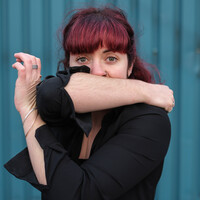 Image of a body in the frame with pink hair. They cover their mouth with their right arm and cross their left arm across their chest so their elbow and left hand connect.