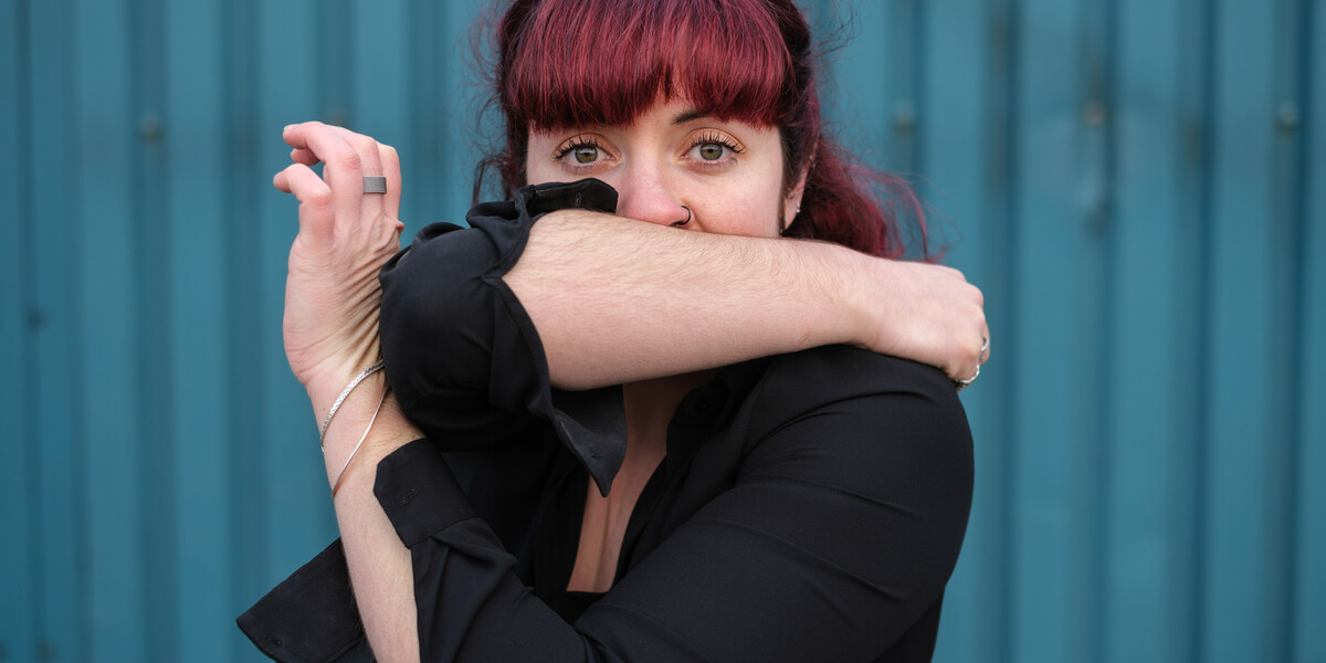 Image of a body in the frame with pink hair. They cover their mouth with their right arm and cross their left arm across their chest so their elbow and left hand connect.